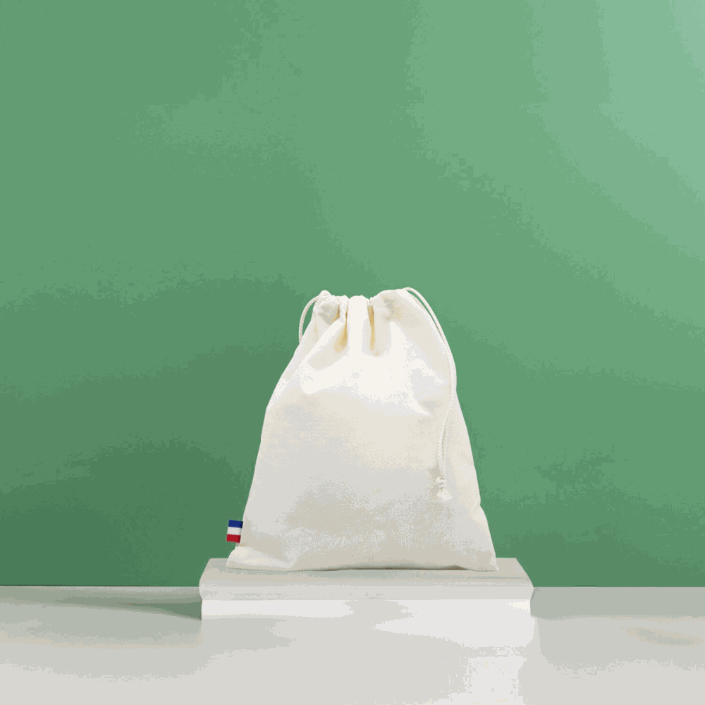 Sac de plage en coton biologique GOTS, grand format, posé sur une serviette au bord de la mer avec accessoires de plage (lunettes, livre, crème solaire)