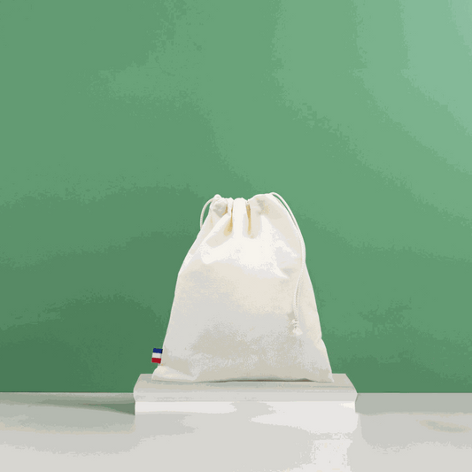 Sac de plage en coton biologique GOTS, grand format, posé sur une serviette au bord de la mer avec accessoires de plage (lunettes, livre, crème solaire)

